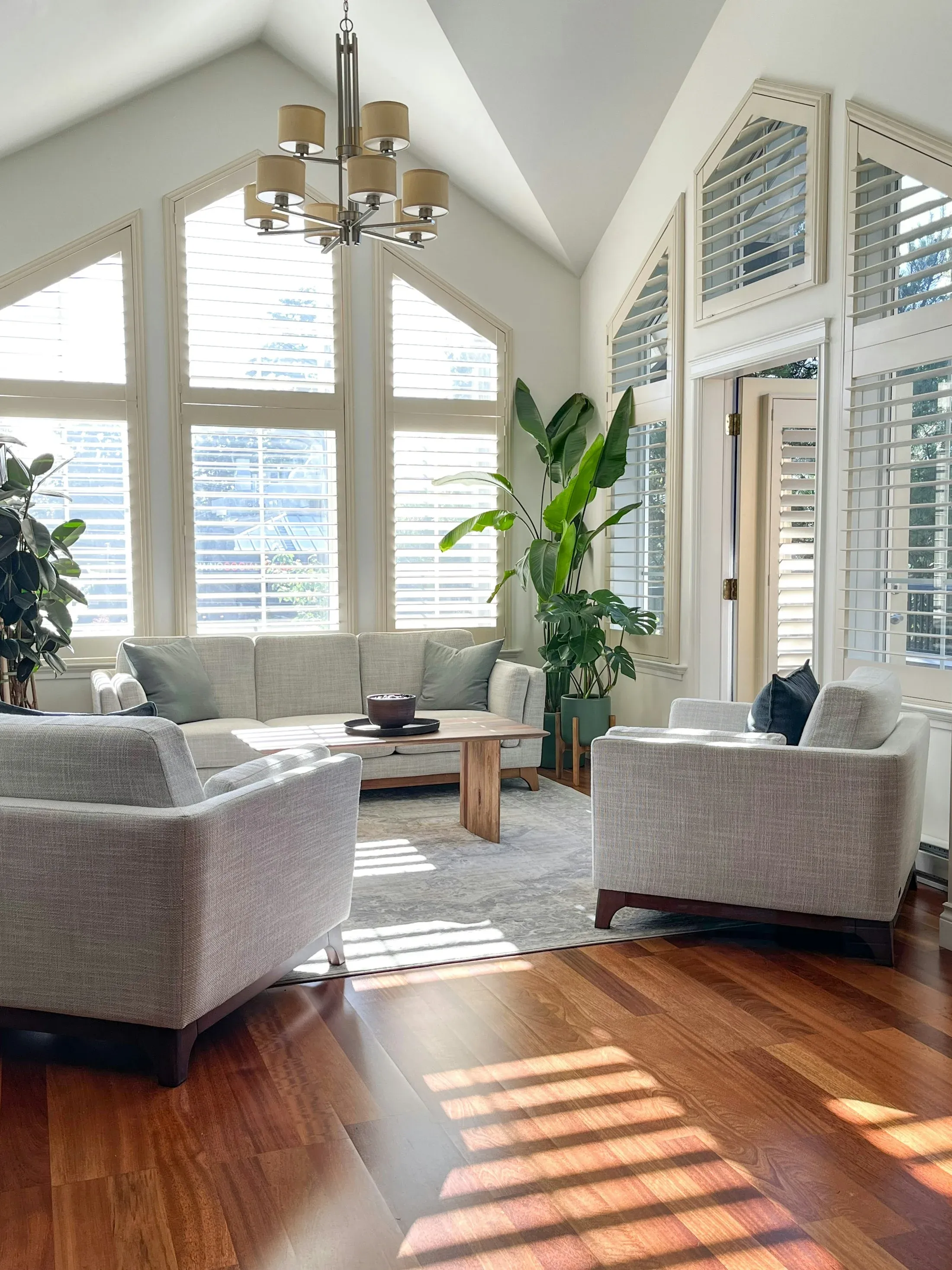 A sunlit sitting room with pale furniture, houseplants, and tall windows